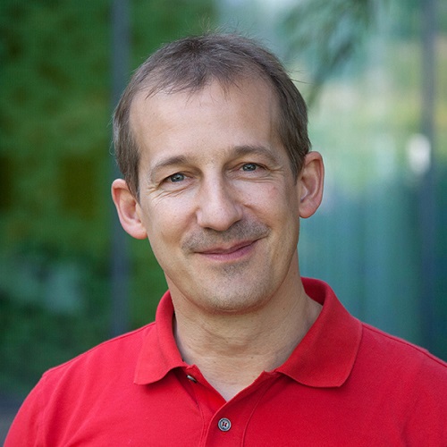 Raimund is a white European man with blue eyes, medium blonde, partly grey hair wearing a red polo shirt. He is directly looking into the camera and smiling kindly.