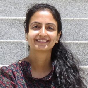 The picture is a headshot of Anupriya Tuli from IIIT-Delhi, India, serving as an assistant to Equity and Justice Chairs, CHI 2022. She is a South Asian female with black eyes and waist-long black curly hair tied into a (left) side ponytail. She is wearing pearl studs and a navy blue printed dress with tiny red and green flowers. She is sitting on light grey stairs (filling the background) with a big friendly smile and dimples on both her cheeks.