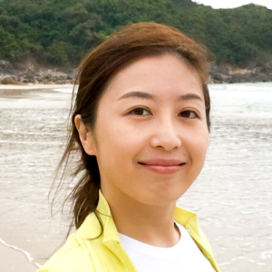 Can Liu smiling towards the camera with the background of water on a beach in front of a green hill.