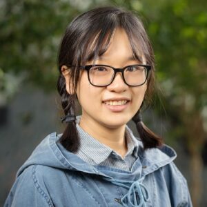 Bingjie Yu is in front of green-leaf plants indoor. She wears glasses with black frame and smiles at the camera.